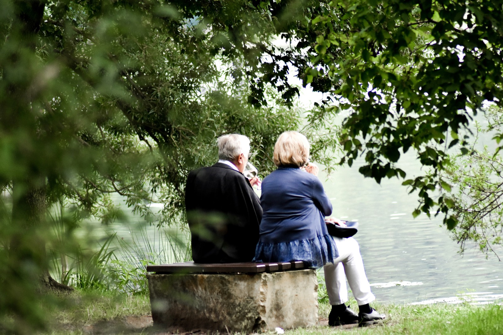 An older couple sitting together on a bench by a lake, sharing a quiet drink